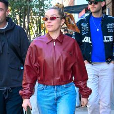 Florence Pugh walks down the street wearing a red leather bomber jacket with blue cuffed jeans and black boots.