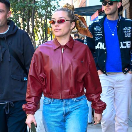 Florence Pugh walks down the street wearing a red leather bomber jacket with blue cuffed jeans and black boots.