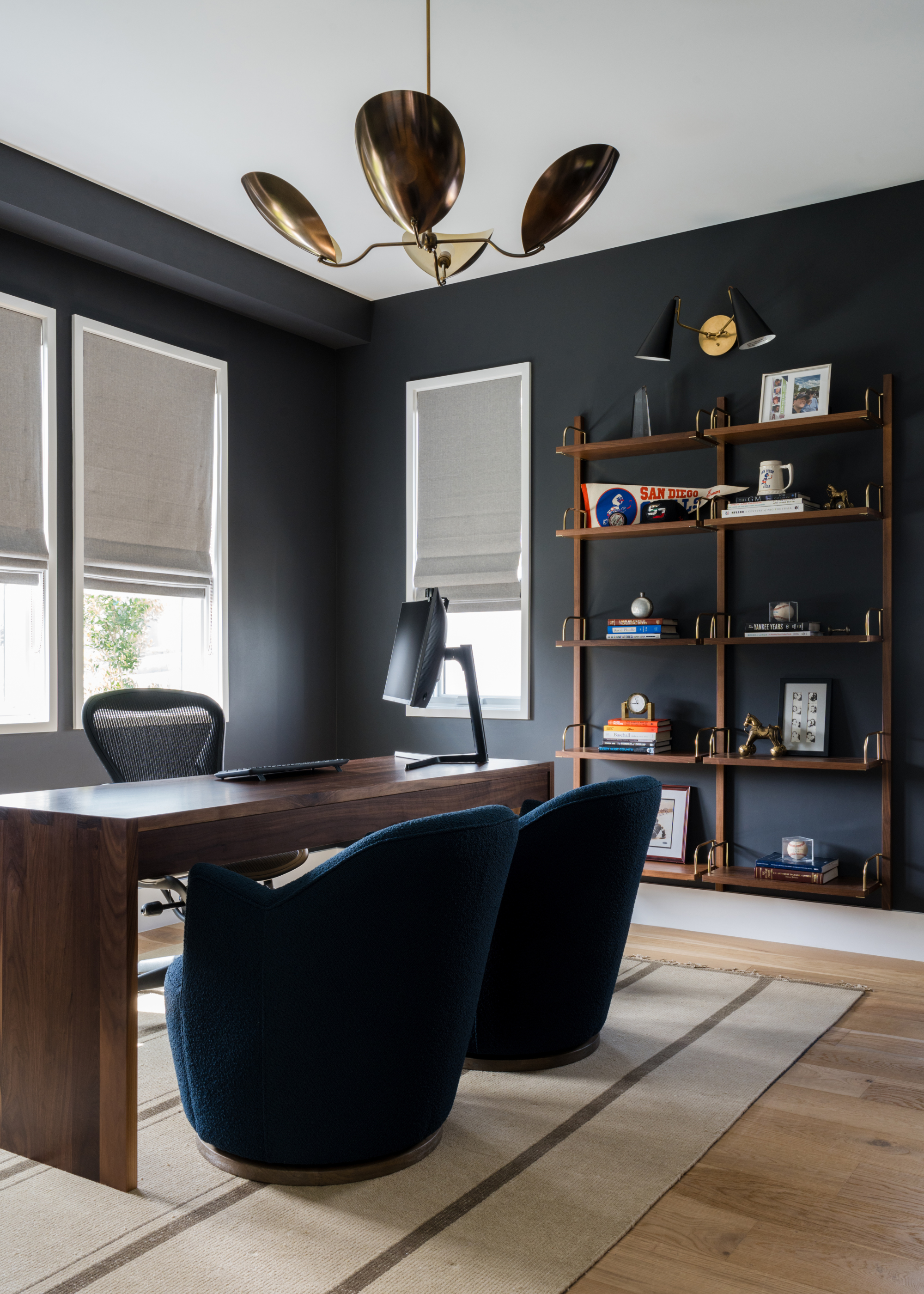 A dark gray home office with a wood desk, a swivel chair, a pair of dark blue armchairs, a striped rug, a wooden shelf with books, prints, and decor