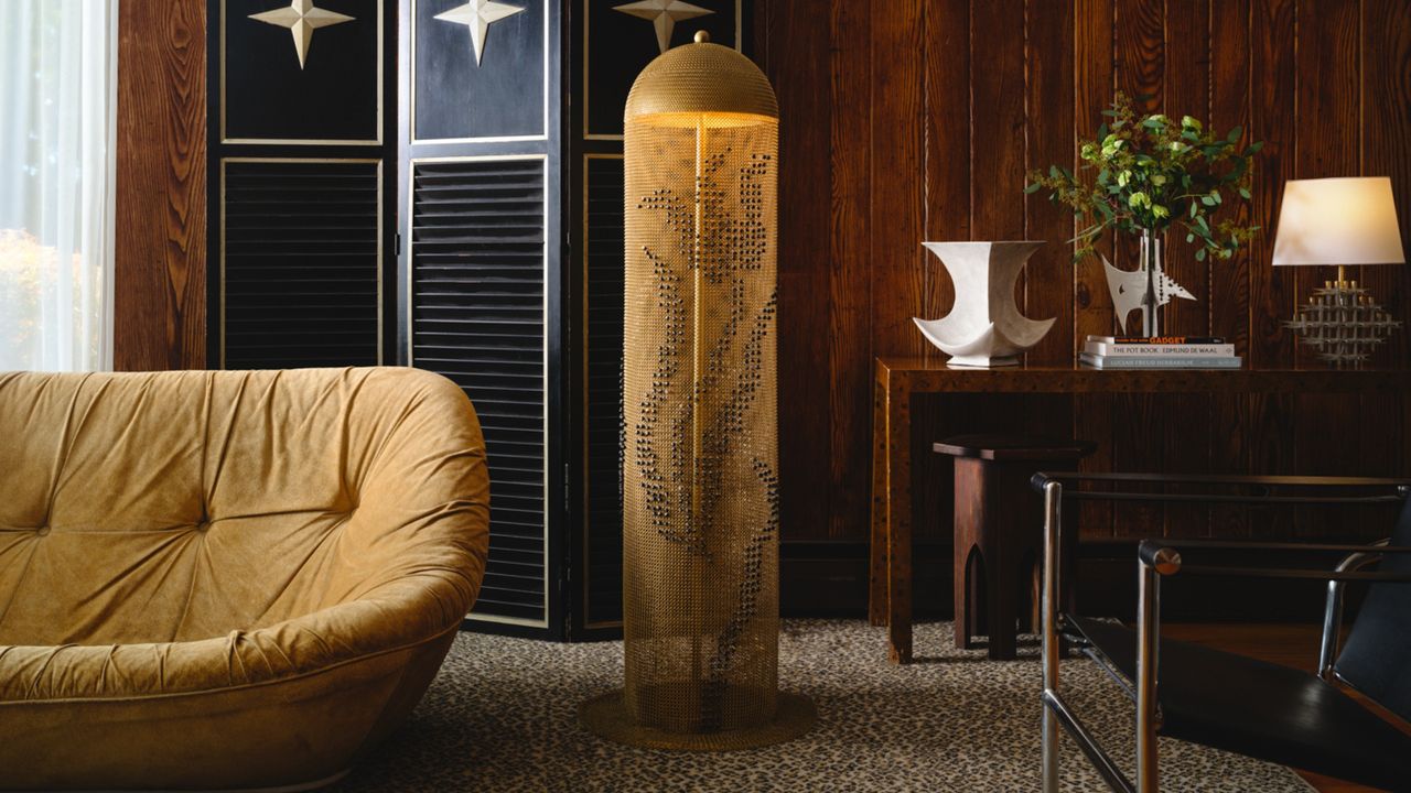 a moody living room design. The walls are dark wood paneling, and there is a wooden desk. On the desk is a white vase, a small plant, and a metal lamp. There is a navy room divider against the wall with a floor lamp in front of it that is draped in chainmail embroidered with beading. Beside the lamp is a dusty yellow loveseat.