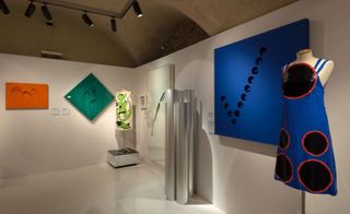 An exhibition space with white walls and floor, featuring abstract painting on the walls in orange, green and blue. Focus on 2 shift dresses by Germana Marucelli on display, one on the left corner and the other on the right corner