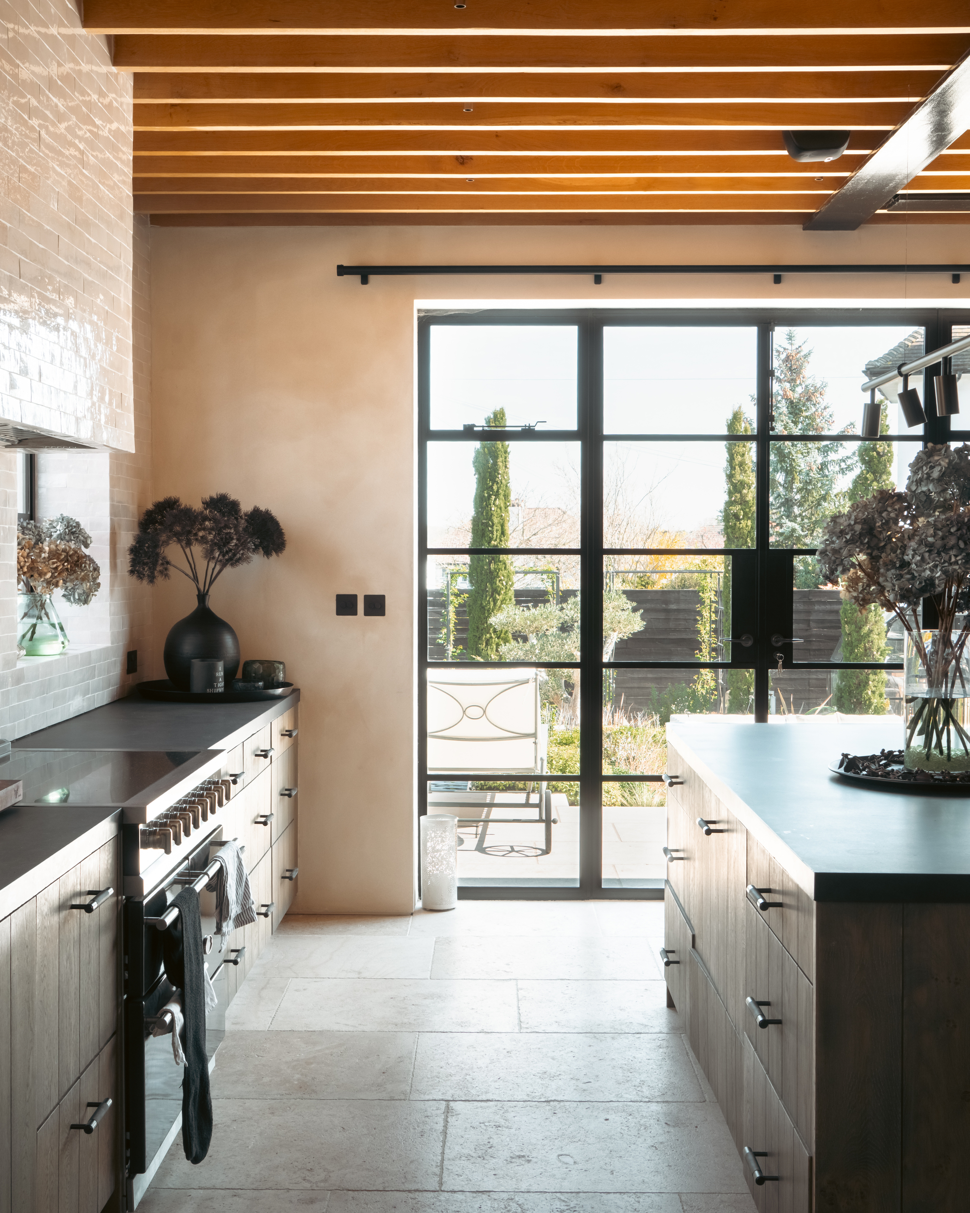 A modern kitchen with limewash walls and gray wooden cabinetry with black countertops. There is a large island and the back wall has large mirrored doors that open to a backyard.