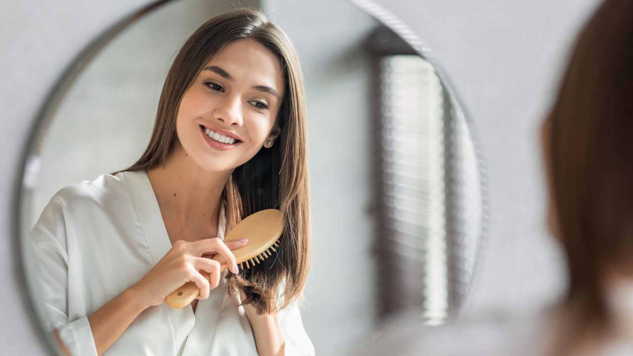 best hair brush - woman smiling in the mirror brushing her hair - gettyimages 1149196482