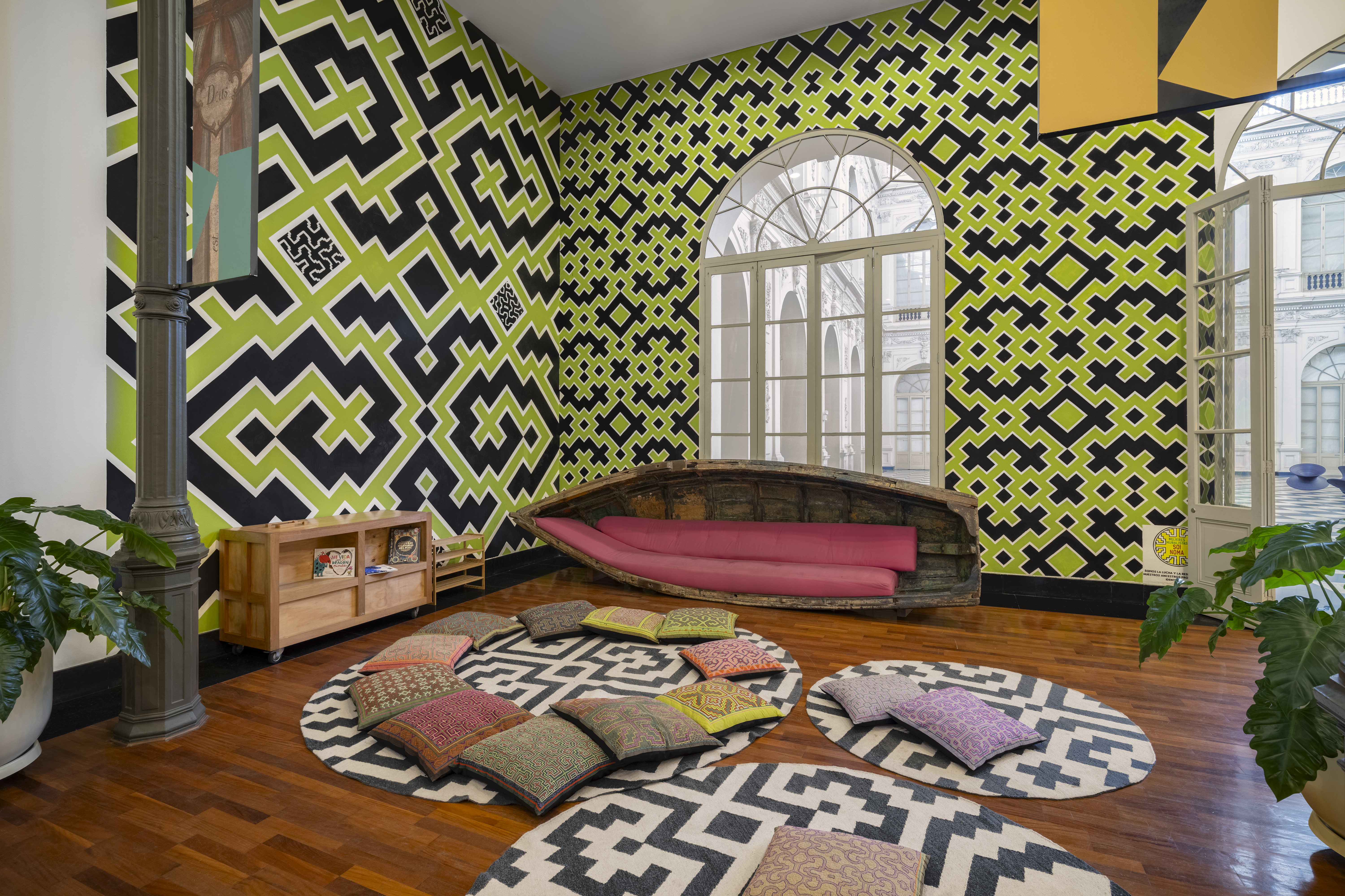 A gallery room with patterned, lime green and blue walls, wooden floors, circular rugs put on the floor and topped with cushions, and thriving plants.