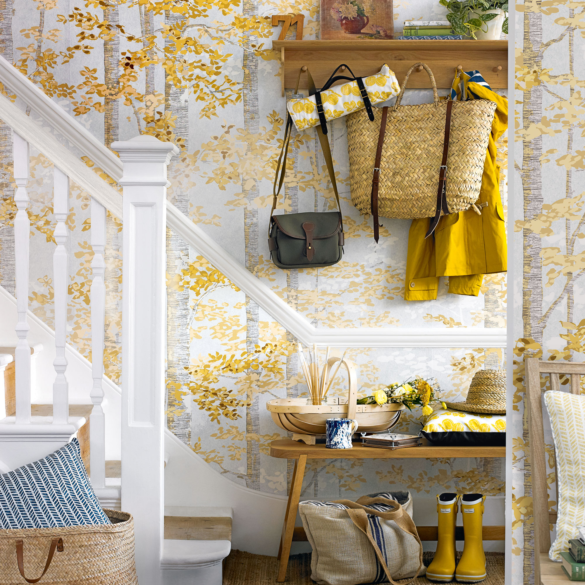 Hallway with yellow tree wallpaper, white staircase, wooden bench and wooden shelf with hooks hanging bags and coats from it