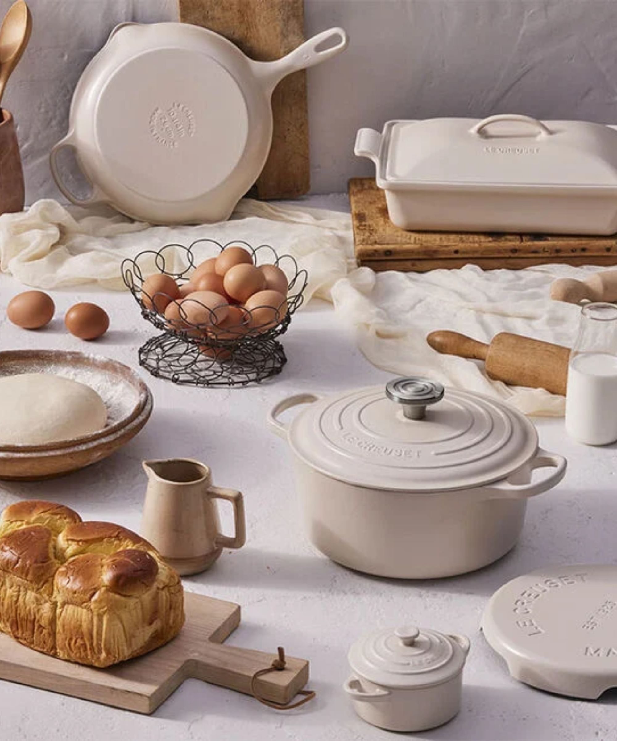 A cream Le Creuset Dutch Oven on a table surrounded by baking essentials, a brioche loaf, and a bown of eggs.