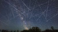 This depicts the satellite-filled sky that is now a reality and getting more crowded every week! This adds together exposures taken over just 30 minutes on an early June night when, from my latitude of 51&deg; N satellites even in low Earth orbit are lit all night by sunlight. Many of the parallel streaks heading generally horizontal west to east (right to left) may be from groups of SpaceX Starlinks. Others traveling vertically north-south are more likely from Earth observation satellites. There is at least one natural streak in the image &mdash; a meteor at centre, caught by chance on one frame. It appears as a colored and tapered streak. Other uniform undashed streaks may be from high-altitude satellites moving much more slowly. By comparison, most satellites appear as dashed lines because the image is a blend of many 2-second-long exposures with a gap of one second between exposures when the camera shutter was closed. So the motion of the satellites and image stacking turns them into dashes. The longer the dashes, the faster the satellite is traveling, with the fastest satellites being the lowest. This is looking due south and all the trails disappear low in the south above the trees, as that's where the Earth's shadow is, even on this June night. So the satellites aren't lit when they are in that small part of the sky. They emerge from the shadow heading north and disappear into the shadow heading south. The shadow creates the obvious boundary of where satellite trails are visible. At other times of the year low-orbit satellites are visible only after sunset or before sunrise, especially from lower latitudes. But not near summer solstice, and from higher latitudes. The field of view is about 100&deg; by 75&deg;. (Photo by: Alan Dyer/VWPics/Universal Images Group via Getty Images)