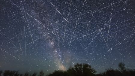 This depicts the satellite-filled sky that is now a reality and getting more crowded every week! This adds together exposures taken over just 30 minutes on an early June night when, from my latitude of 51&deg; N satellites even in low Earth orbit are lit all night by sunlight. Many of the parallel streaks heading generally horizontal west to east (right to left) may be from groups of SpaceX Starlinks. Others traveling vertically north-south are more likely from Earth observation satellites. There is at least one natural streak in the image &mdash; a meteor at centre, caught by chance on one frame. It appears as a colored and tapered streak. Other uniform undashed streaks may be from high-altitude satellites moving much more slowly. By comparison, most satellites appear as dashed lines because the image is a blend of many 2-second-long exposures with a gap of one second between exposures when the camera shutter was closed. So the motion of the satellites and image stacking turns them into dashes. The longer the dashes, the faster the satellite is traveling, with the fastest satellites being the lowest. This is looking due south and all the trails disappear low in the south above the trees, as that's where the Earth's shadow is, even on this June night. So the satellites aren't lit when they are in that small part of the sky. They emerge from the shadow heading north and disappear into the shadow heading south. The shadow creates the obvious boundary of where satellite trails are visible. At other times of the year low-orbit satellites are visible only after sunset or before sunrise, especially from lower latitudes. But not near summer solstice, and from higher latitudes. The field of view is about 100&deg; by 75&deg;. (Photo by: Alan Dyer/VWPics/Universal Images Group via Getty Images)