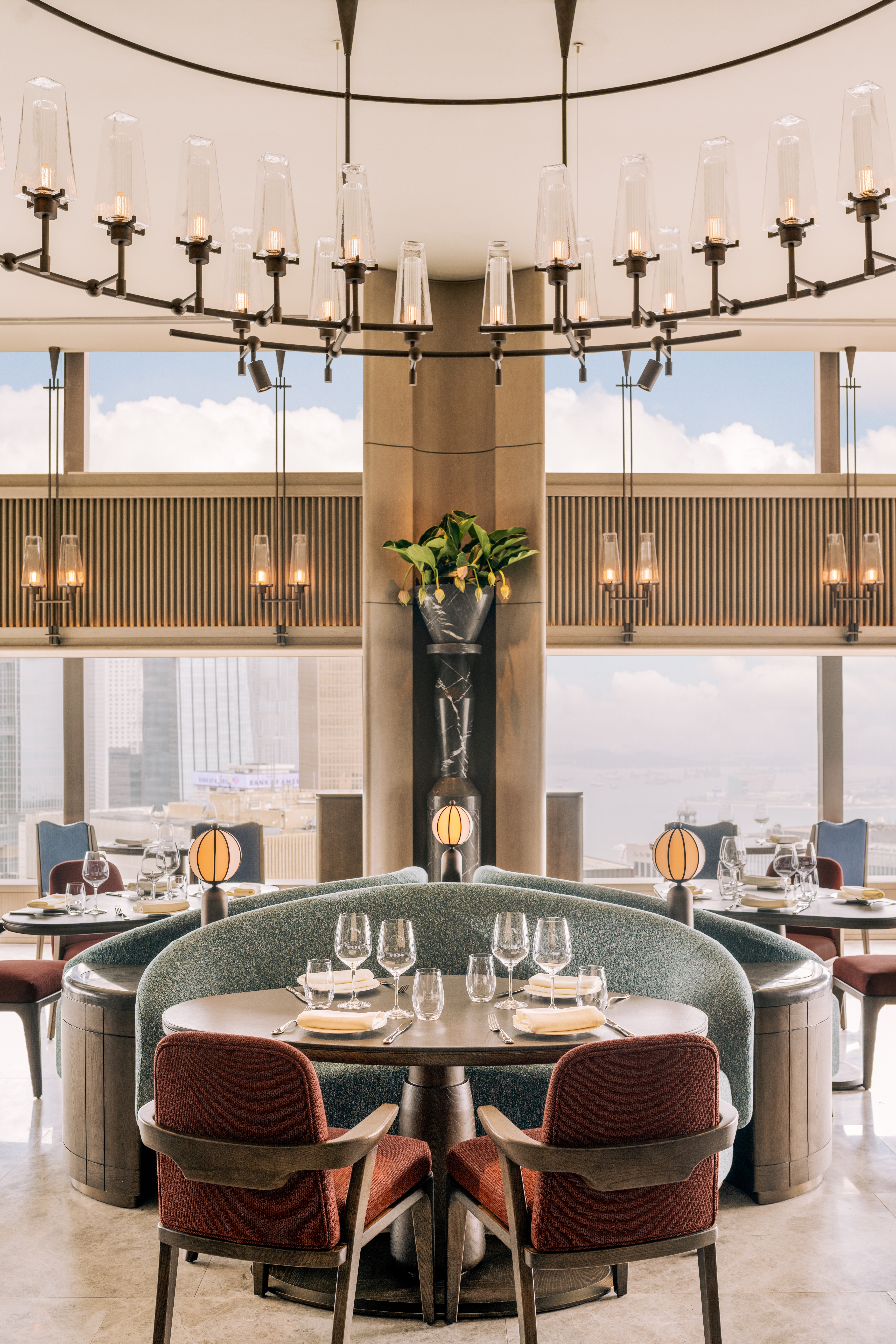 A glamorous bar situated on the top floor of a hotel, with views over the surroundings skyscrapers, is decorated with vintage-inspired, sculptural banquettes in seafood green and brick red chairs, glass sconces and chandeliers, and air balloon-shaped table lamps.