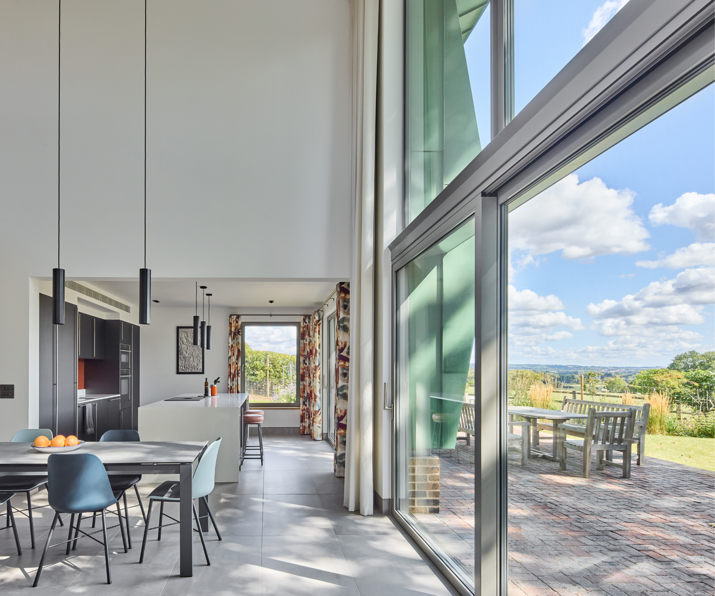 Modern open plan kitchen and dining area with large floor to ceiling windows and patio doors overlooking garden and brick patio with seating