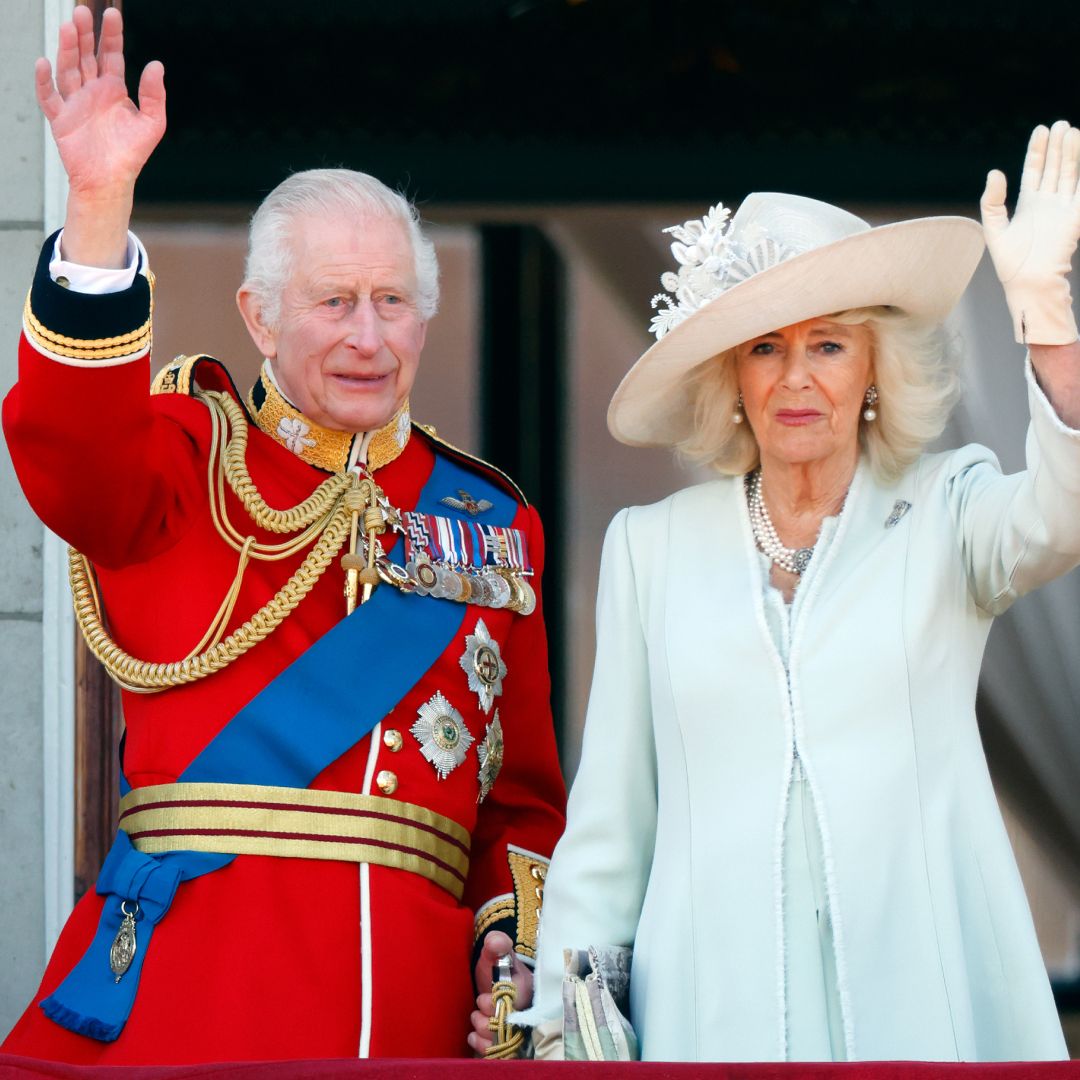 King Charles wearing a red and gold military uniform waving and smiling standing next to Queen Camilla in a mint coat dress also waving and smiling 