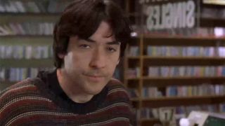 John Cusack in a sweater, looking annoyed and looking into the camera in High Fidelity