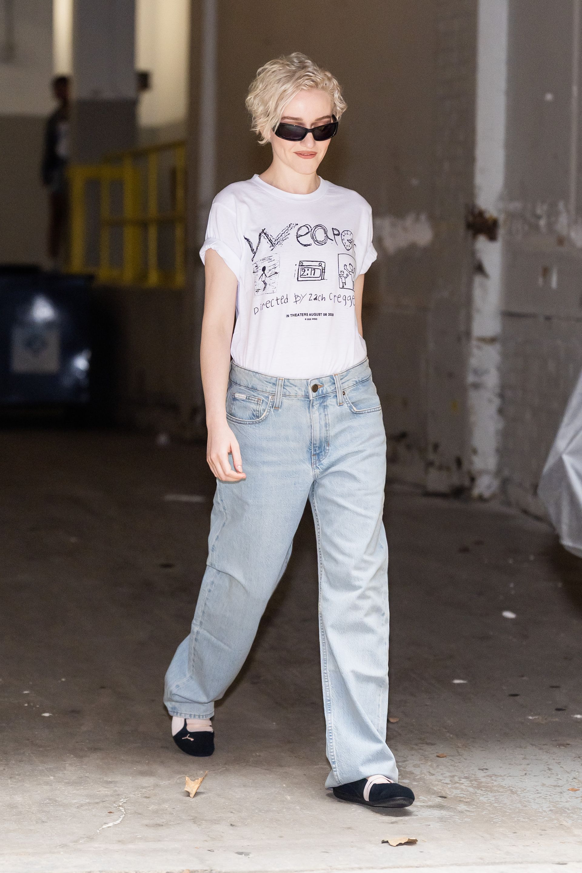 Julia Garner wearing a white graphic T-shirt with baggy jeans and black Puma Speedcat sneakers