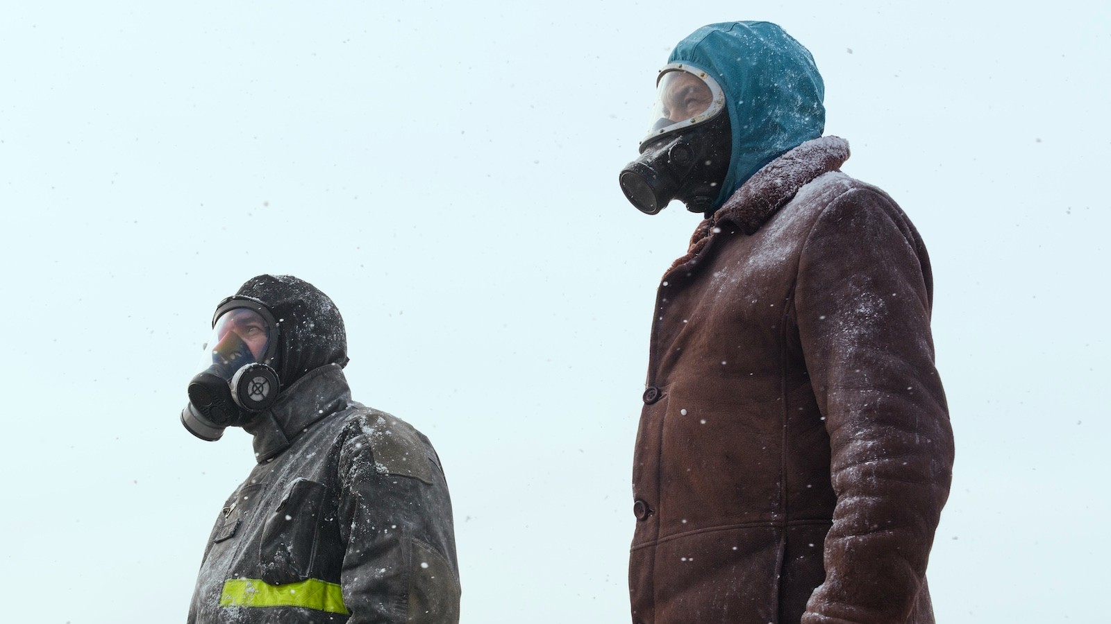 Scene from El Eternauta: Two individuals, both wearing gas masks and winter clothing, stand outdoors in a snowy, overcast environment, looking into the distance.
