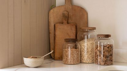 A set of three larhe glass storage jars with wooden lids in front of two wooden cutting boards and next to an stonewarebowl of cereal
