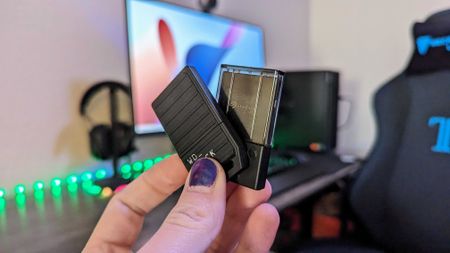 A hand holding a small, black electronic device in a gaming setup, with a colorful, blurred monitor and gaming chair in the background.