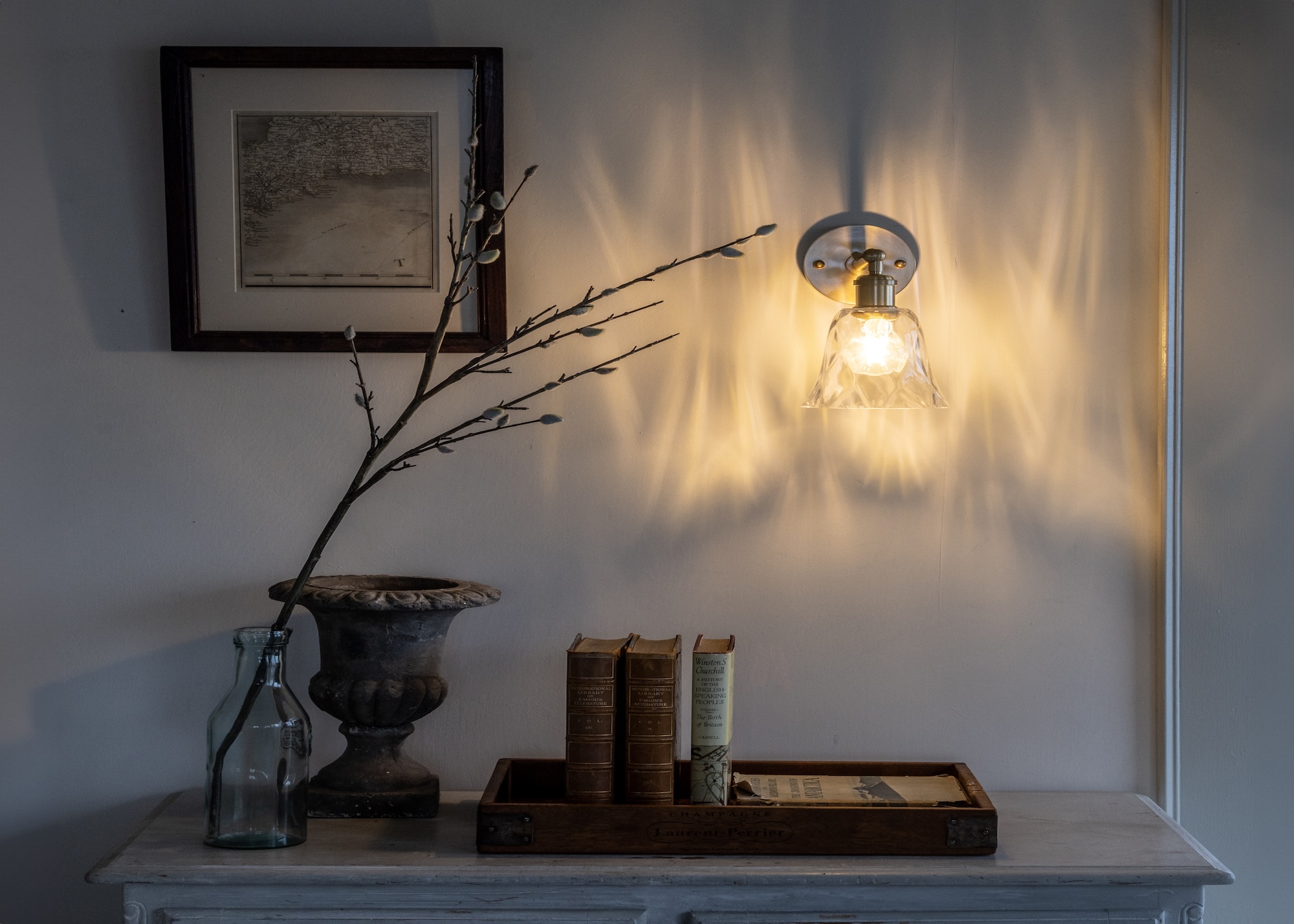 An entryway with a bright light shining above an entryway table. On the table is a tray with books standing upright as well as a glass base with a stem inside of it.