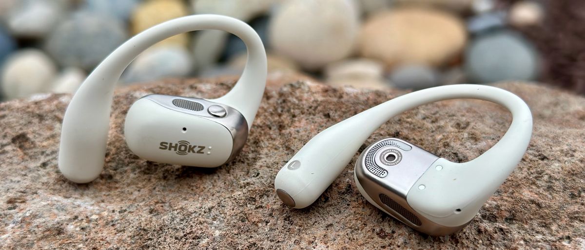 A close-up of the Shokz OpenFit 2 earbuds, one facing the camera showing the logo and top button/speaker grill, the other laying on its back showing the main speaker grill, mic hole, and ear hook.