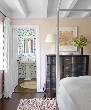 bedroom with pale pink walls, white four poster, wooden chest of drawers with ensuite with green patterned wallpaper