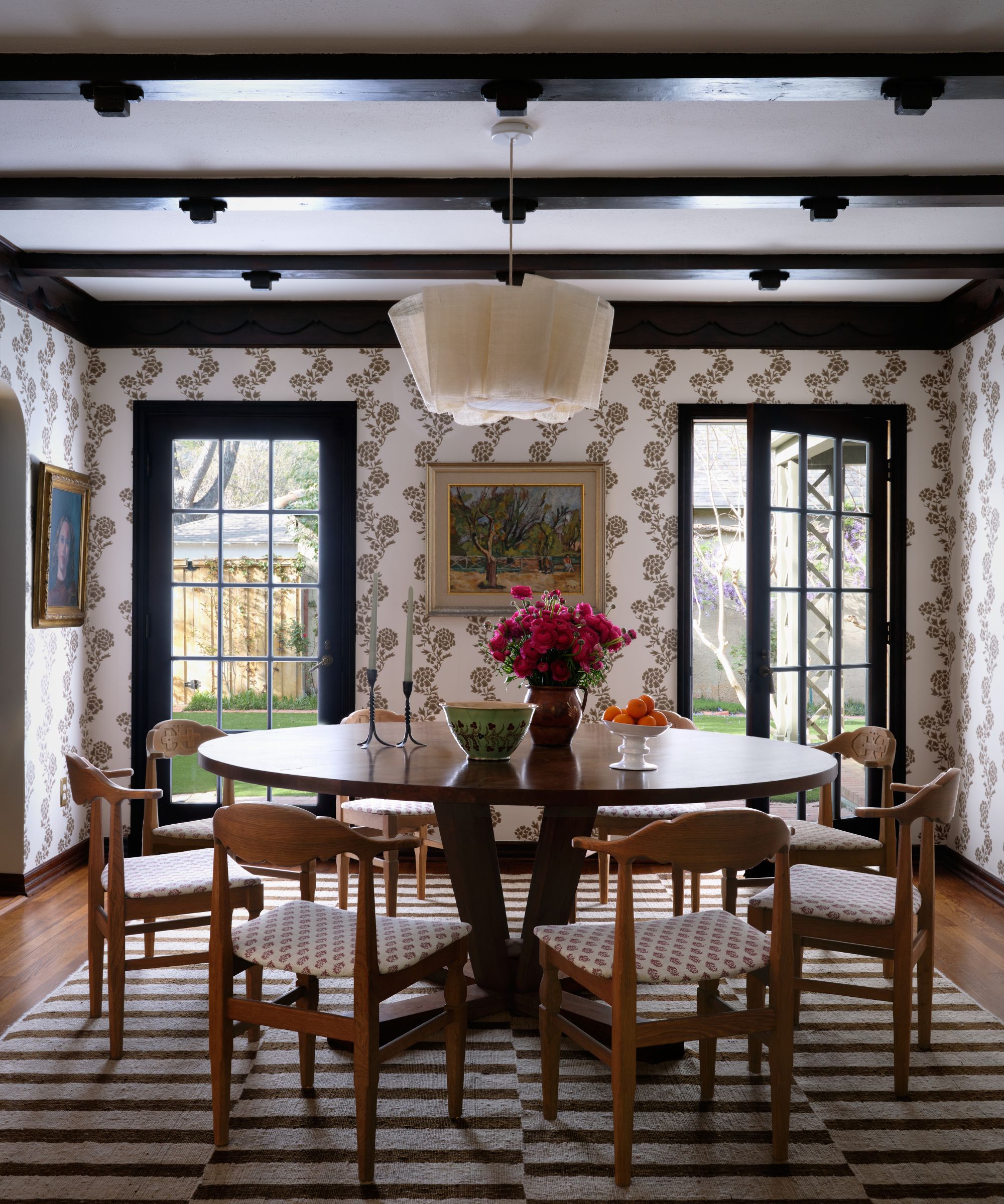 A wide view of a dining room showing a round wooden table under a linen pendant light, surrounded by dark wood ceiling beams and French doors