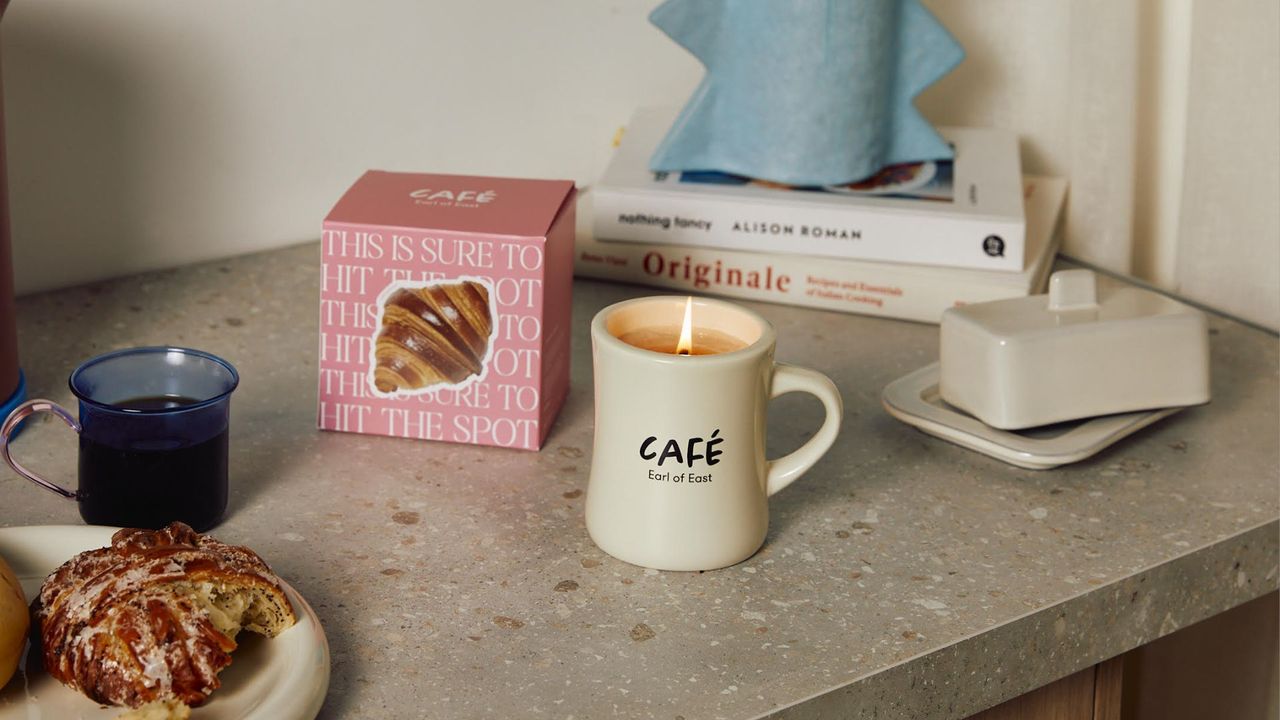 A terrazzo countertop with a couple of books, a textured blue vase, a mug of coffee, a glazed croissant on a plate, a butter dish and a mug candle alongside its boxed packaging.