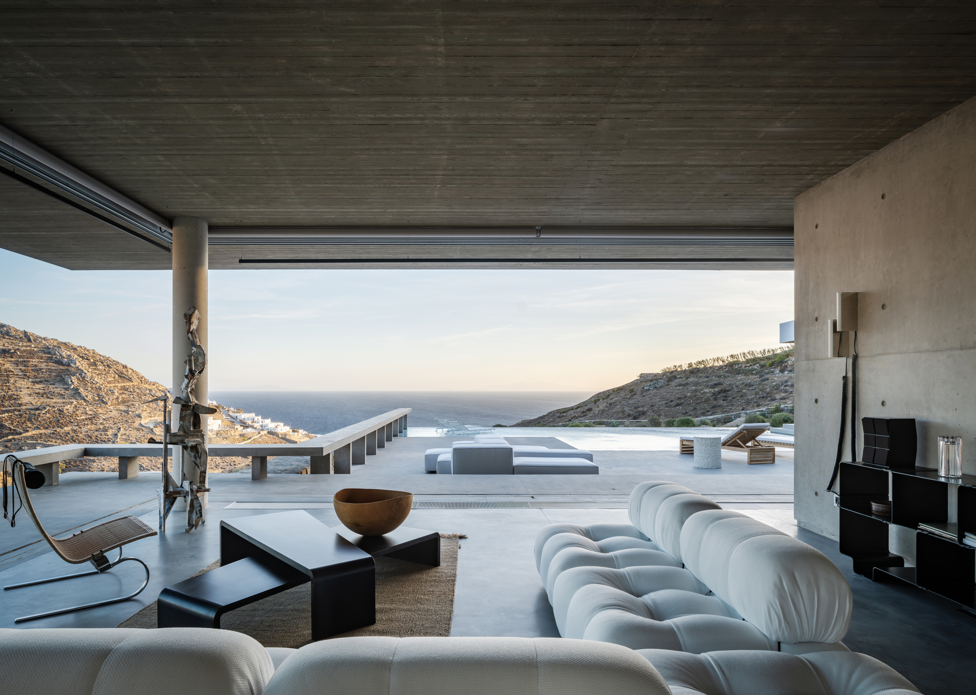 modern living room with iconic designer furniture, concrete ceiling, floors, and walls, indoor-outdoor connection leading to outdoor area with infinity pool