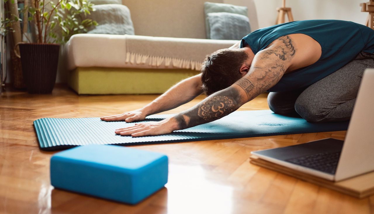 Man doing yoga at home on a blue yoga mat. He is kneeling with his head down and arms stretched out in front of him, palms face down on the mat. A blue yoga block and open laptop are on the floor in the foreground. The man is wearing gray yoga pants and a dark green vest top. He has sleeve tattoos on his left arm. 