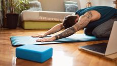 Man doing yoga at home on a blue yoga mat. He is kneeling with his head down and arms stretched out in front of him, palms face down on the mat. A blue yoga block and open laptop are on the floor in the foreground. The man is wearing gray yoga pants and a dark green vest top. He has sleeve tattoos on his left arm. 