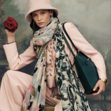 Model wearing a baby pink hat and suit set with patterned scarves and shoulder bag sold at Accessorize a rose in her right hand 