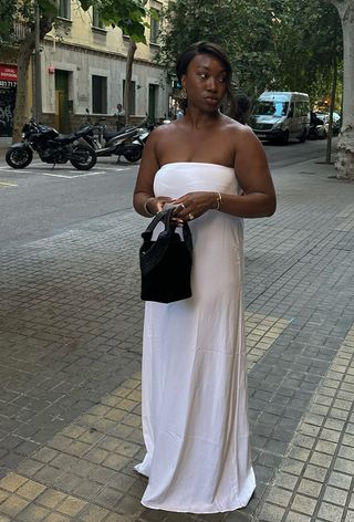 A woman wearing gold sculptural earrings, a white strapless maxi dress, a black bag, and gold bangles.