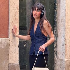 Influencer @tamaramory stand outside wearing a denim halterneck top with black trousers. She carries a white handbag and wears headphones over her ears.