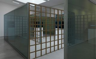 Close up of Installation view of Greek Meander Pavilion, room with slate grey floor, white walls, suspended white ceiling with skylights and spotlights around the edge, wooden frame construction with blue glass panel dividers and white floor