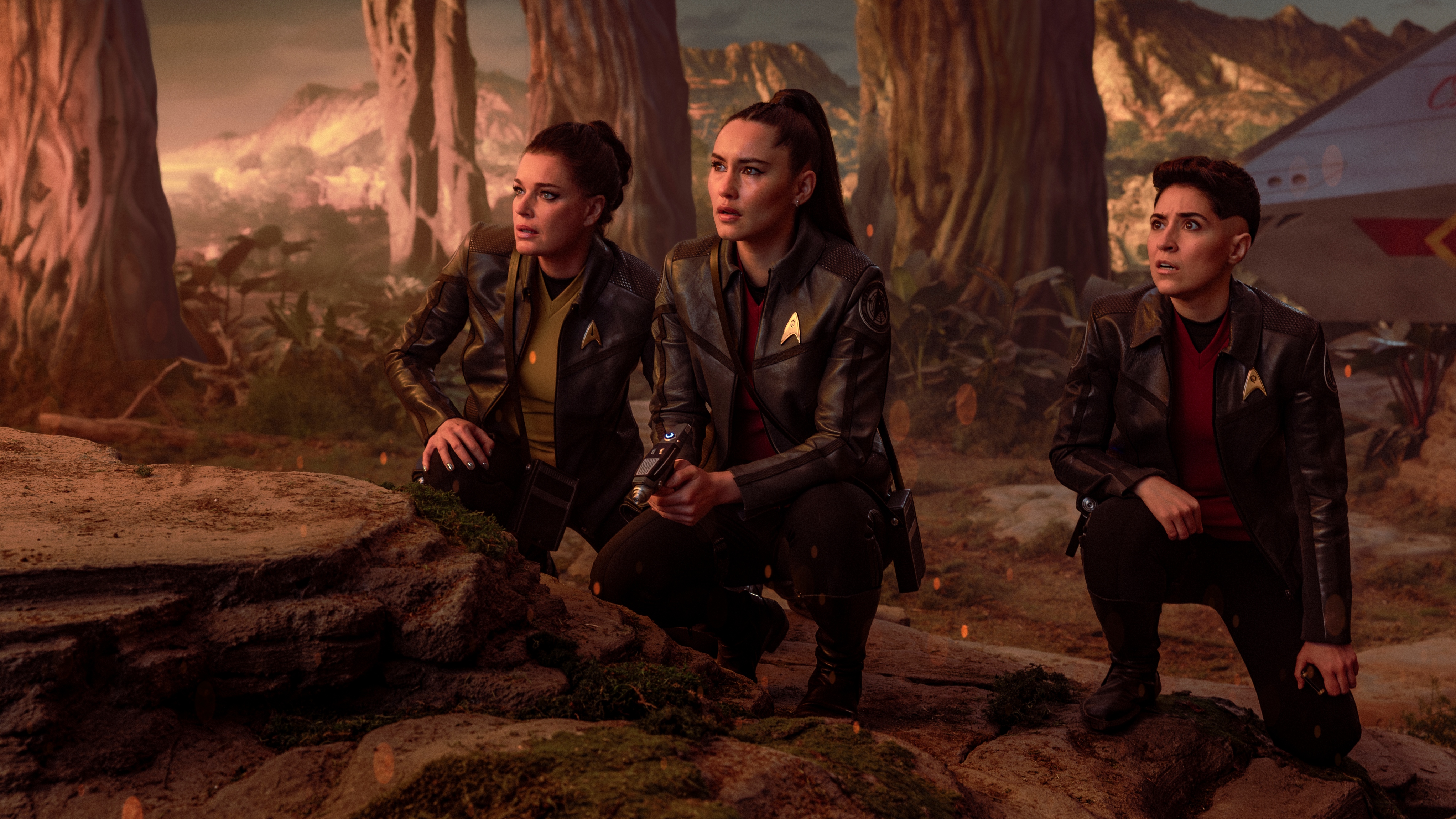 (L-R): Rebecca Romijn as Una, Christina Chong as La&amp;rsquo;an Noonien-Singh, and Melissa Navia as Erica Ortegas crouching on rocks.