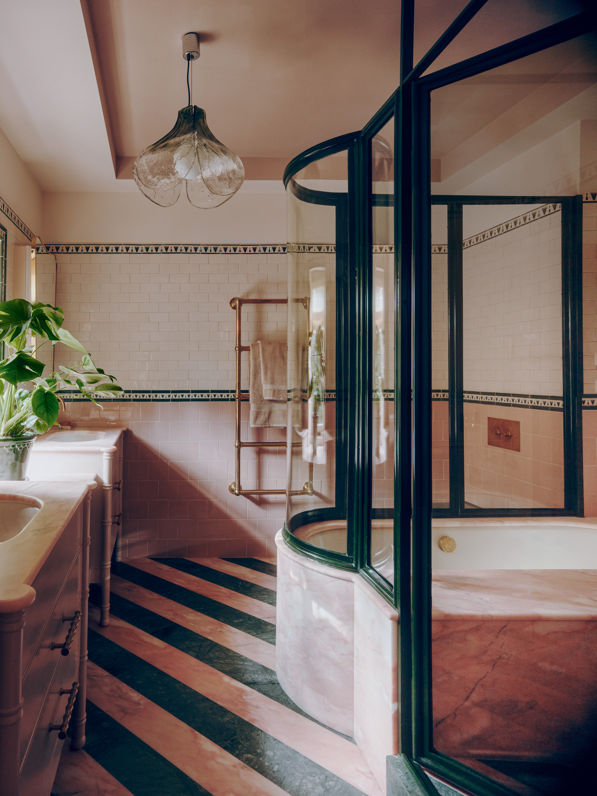 Pink and green wallpaper with striped stone floor, dark green framed glass shower and bath screen