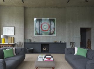A lounge area with grey sofa's, a grey foot stool, a grey console, a fireplace, a TV and concrete walls.