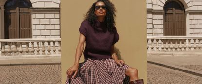 Model wearing a Mint Velvet plum purple knit vest, black sunglasses and a plaid maxi skirt sat in front of a board in an urban street