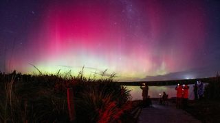 a group of people stand at the edge of a body of water to view bring yellow white and magenta waves of ionized gas in Earth's atmosphere, known as aurora borealis or the northern lights
