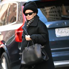 Ashley olsen wears a black hat, black sunglasses, plaid scarf, long black coat, gray socks, black the row margaux bag, and black v2k nike sneakers.