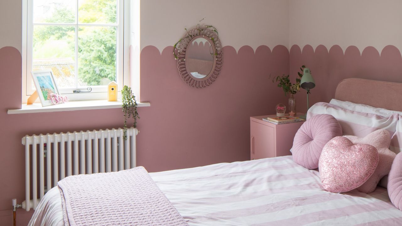 A pink bedroom with a wall divided in half with a scalloped motif and striped bedding on the bed