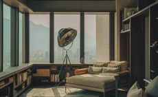 A photo of a penthouse overlooking a city. The furnishings include a low-backed chair with beige cushions, a standing light with a moon-like light shade and large portrait bay windows. 