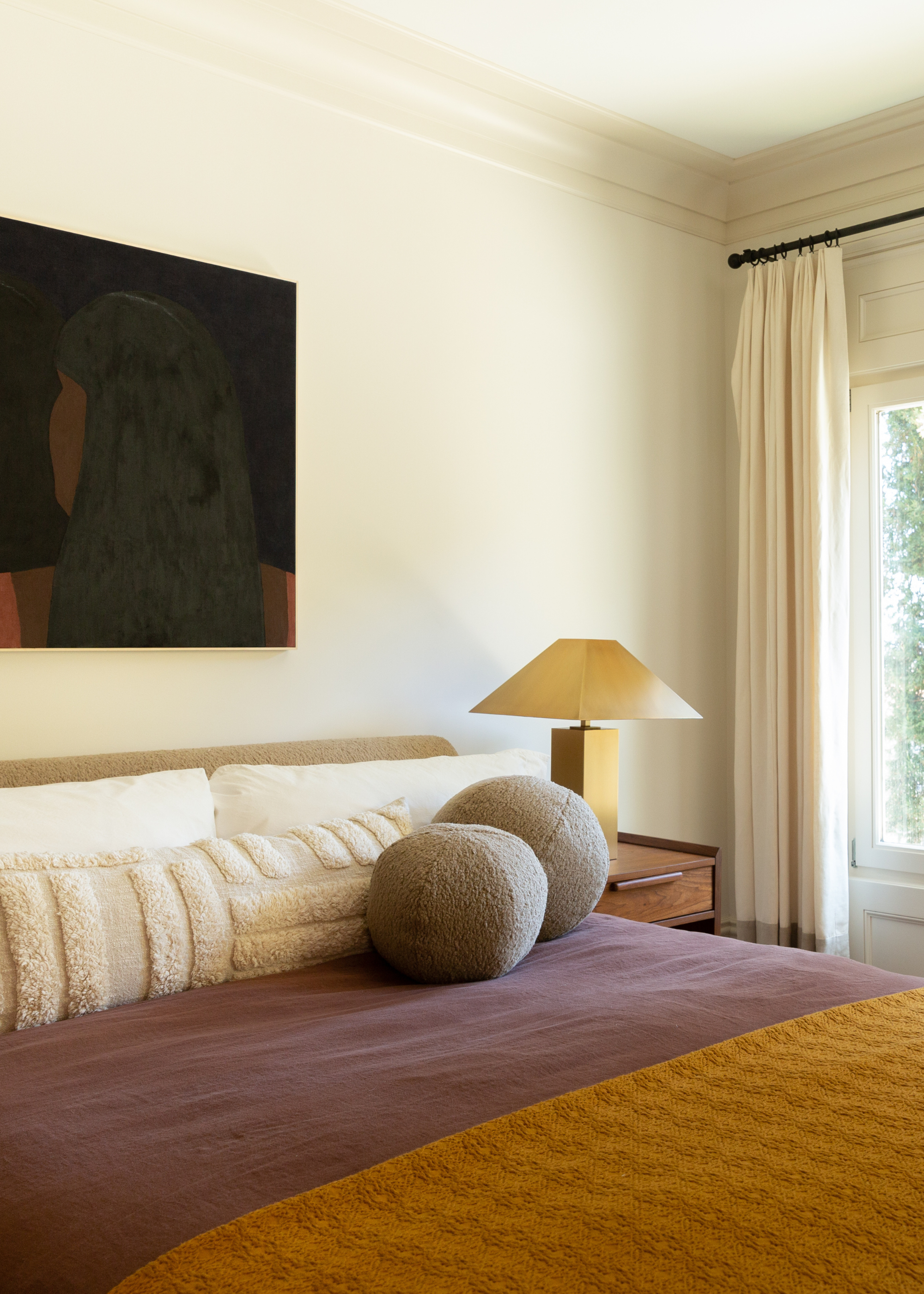 A white bedroom with a dark framed painting, a bed with a textured bolster pillow and a pair of gray fuzzy round pillows on a purple duvet with a burnt yellow throw, beside a wood side table with a gold lamp