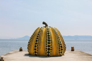 A colossal yellow pumpkin with painted black dots sits on a cement harbor facing a calm seaview.