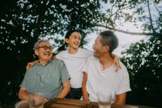 Three generations of a family smile and laugh with each other outside in warm weather.