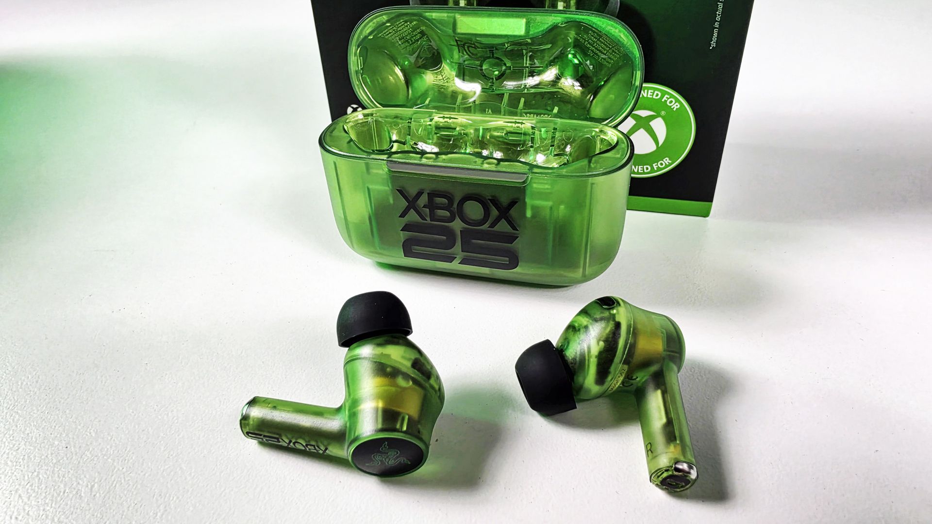 Open translucent green charging case with the Razer Hammerhead V3 X HyperSpeed wireless earbuds sitting in front against white tabletop.