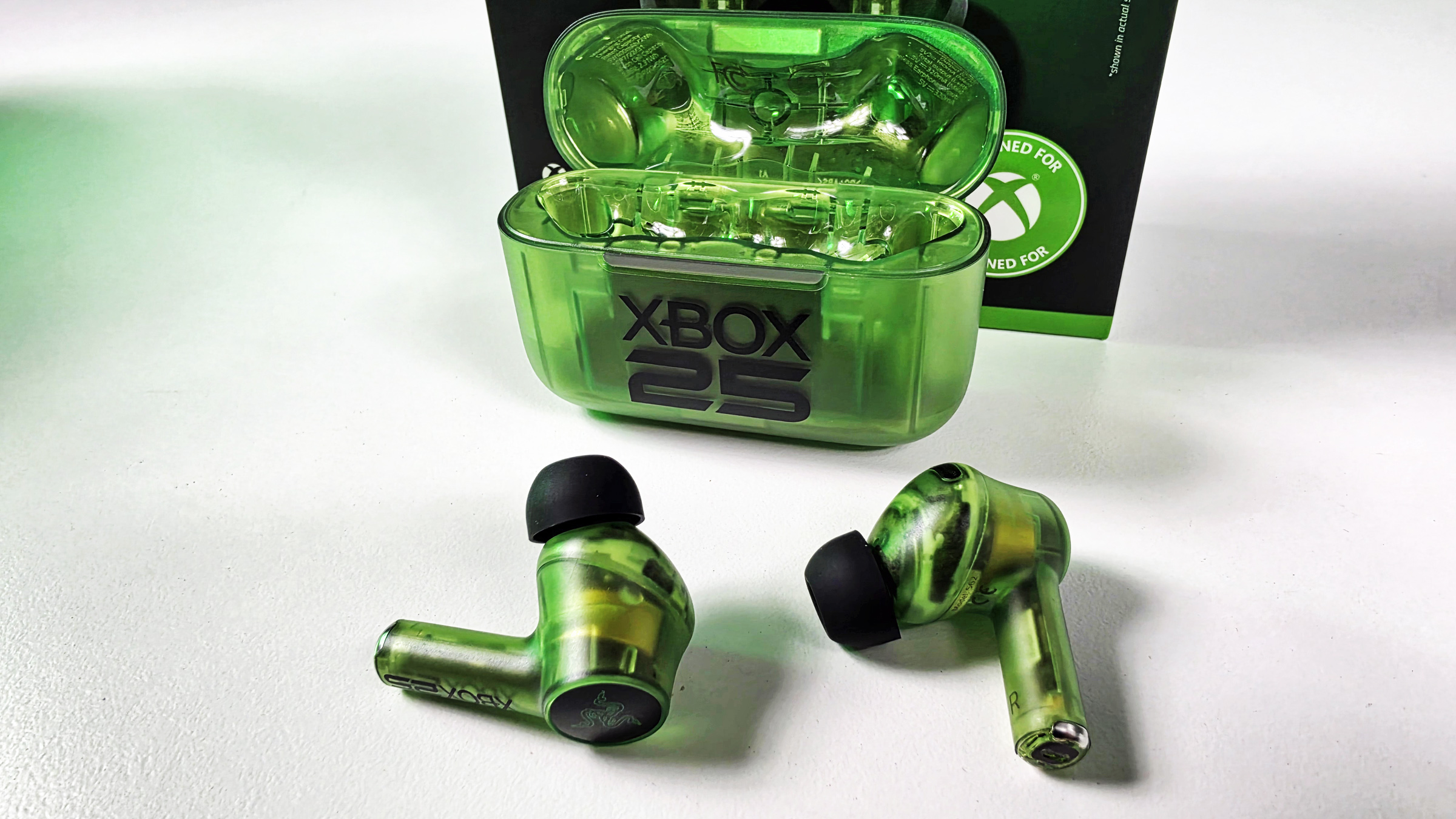 Open translucent green charging case with the Razer Hammerhead V3 X HyperSpeed wireless earbuds sitting in front against white tabletop.