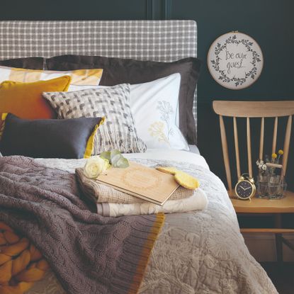 A dark guest bedroom with the bed layered with patterned scatter cushions and throws and a stack of towels on top of the bed