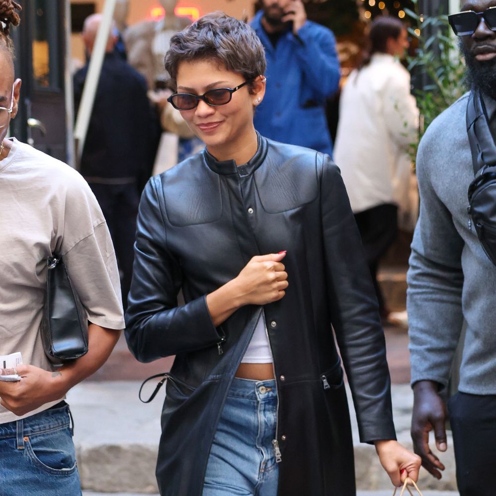 Zendaya steps outside in Paris wearing a black leather coat with blue cuffed jeans, heeled Tabis and black sunglasses. She carries a paper shopping bag.