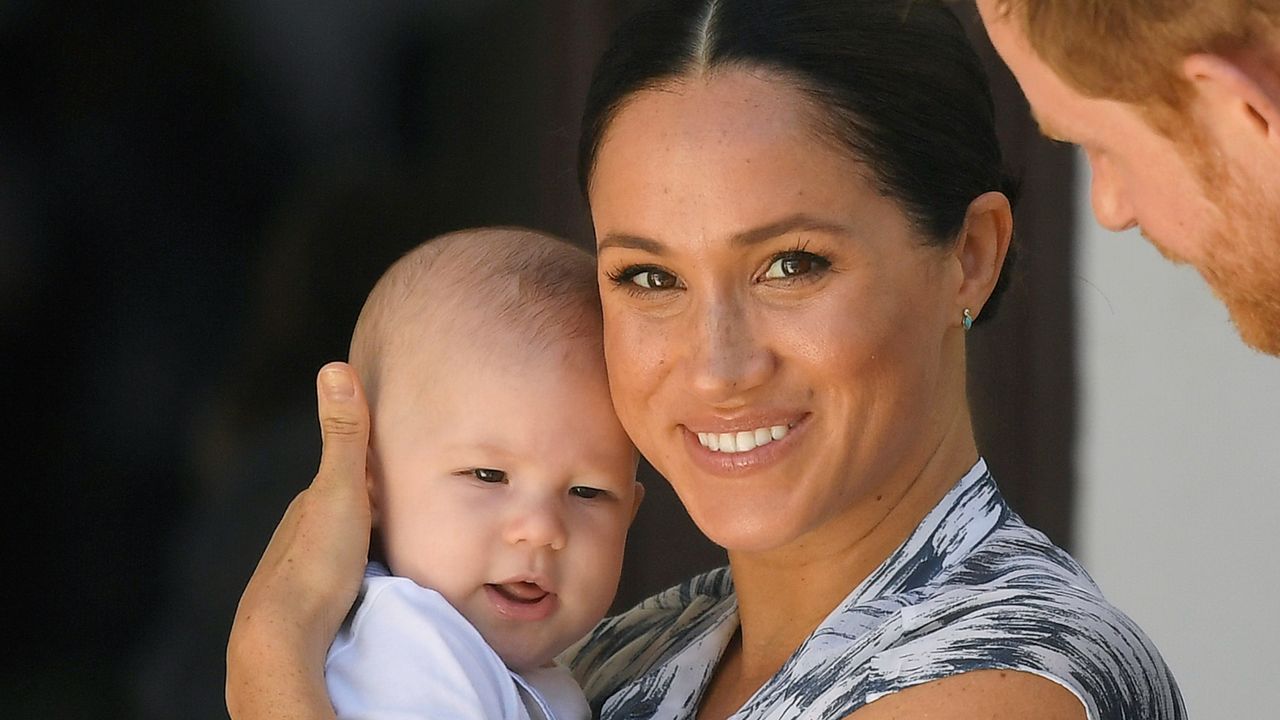 Meghan Markle wears a blue and white dress and cradles a young Prince Archie