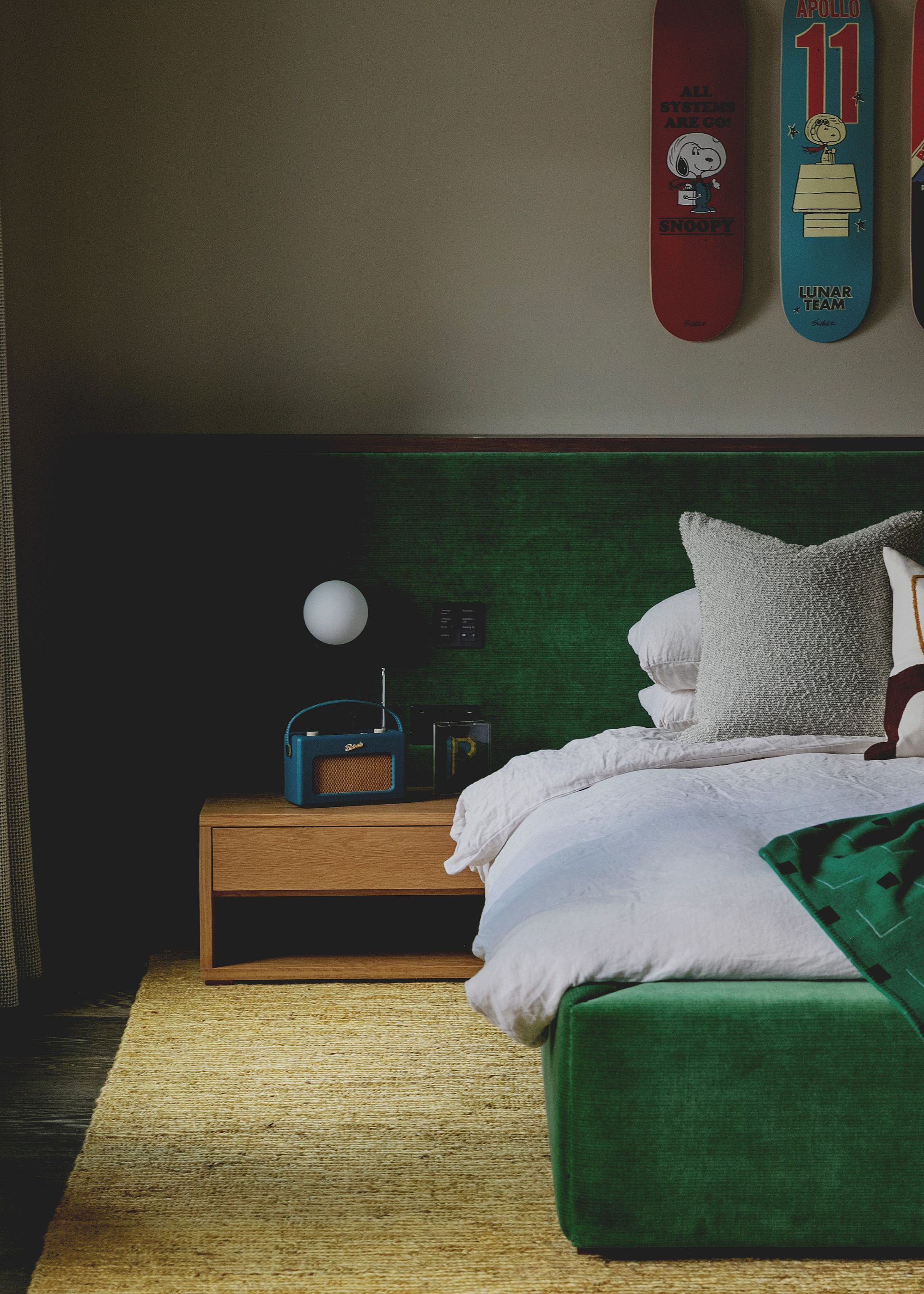 A modern bedroom with a dark green velvet headboard, white bedding, a wooden nightstand, and a light green rug on the floor.