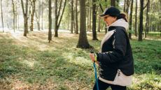 Woman doing metabolic walking holding pole and walking through forest wearing activewear and cap with coat on